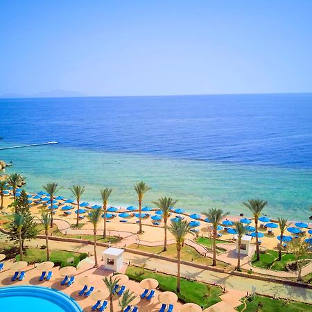 Pickalbatros Royal Grand Sharm - Adults Friendly 16 Years Plus ภายนอก รูปภาพ The photo displays a beautiful coastal scene featuring a clear blue sea that transitions into green tones near the shore. In the foreground, there are sandy beaches lined with palm trees and a variety of beach umbrellas. There is also a swimming pool