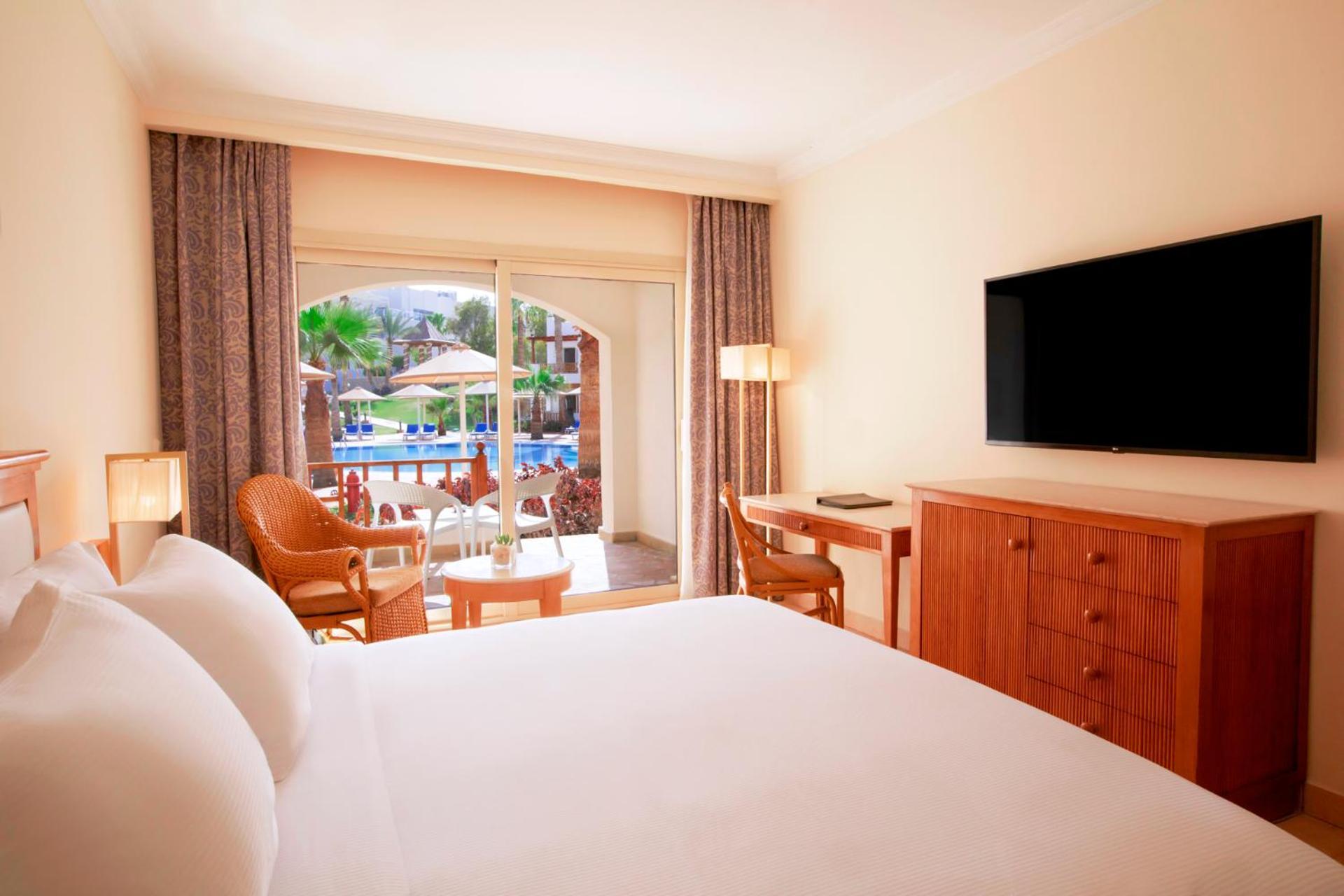 Pickalbatros Royal Grand Sharm - Adults Friendly 16 Years Plus ภายนอก รูปภาพ The photo shows a hotel room featuring a large bed with white bedding. There are wooden furniture pieces, including a dresser and a desk. A flat-screen television is mounted on the wall opposite the bed. The room has large windows with sheer curtains