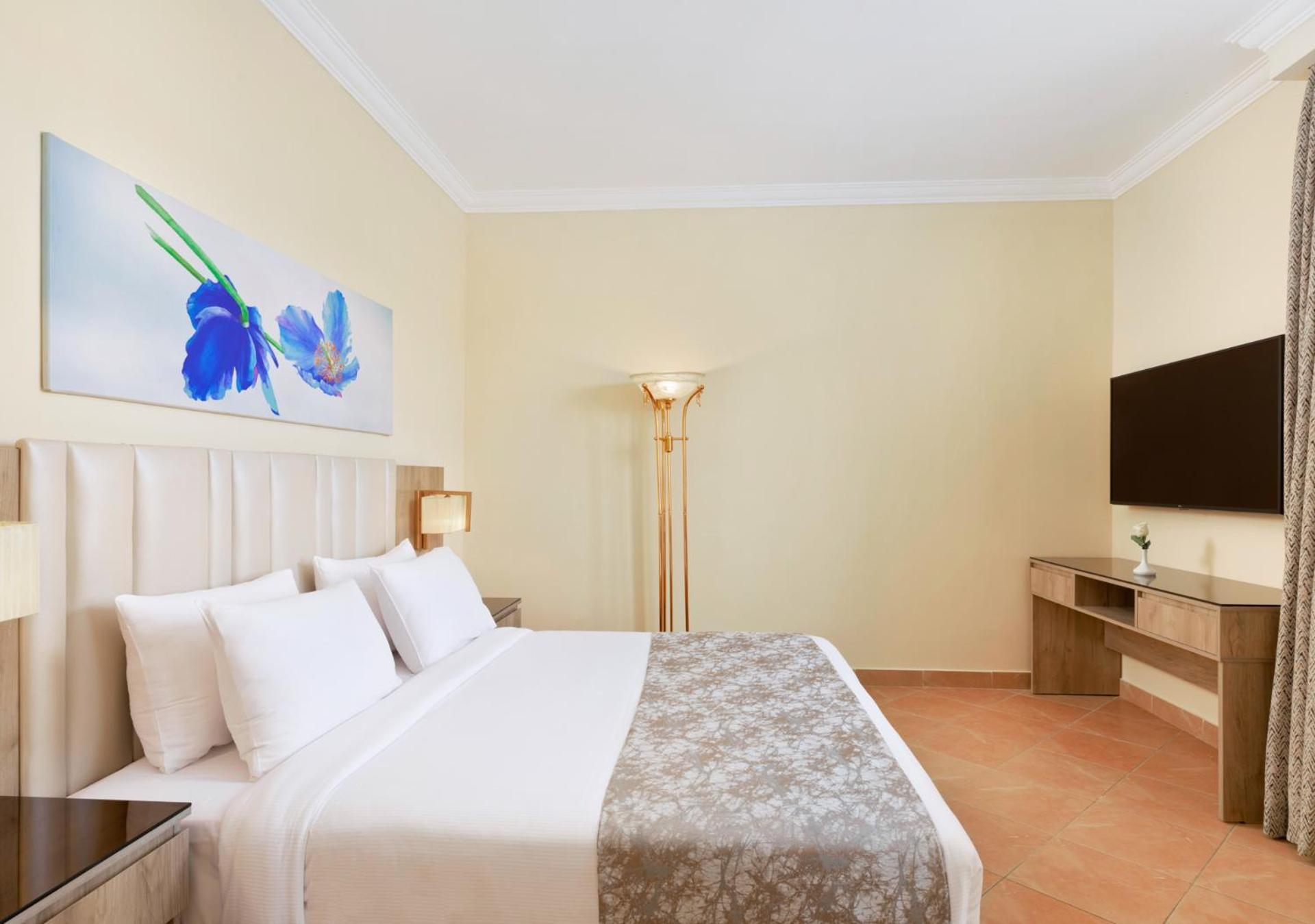 Pickalbatros Royal Grand Sharm - Adults Friendly 16 Years Plus ภายนอก รูปภาพ The photo shows a modern hotel room featuring a large bed with white linens and a decorative gray throw. On one side of the room, there is a piece of artwork depicting blue flowers. A floor lamp provides additional lighting, and across from the bed i