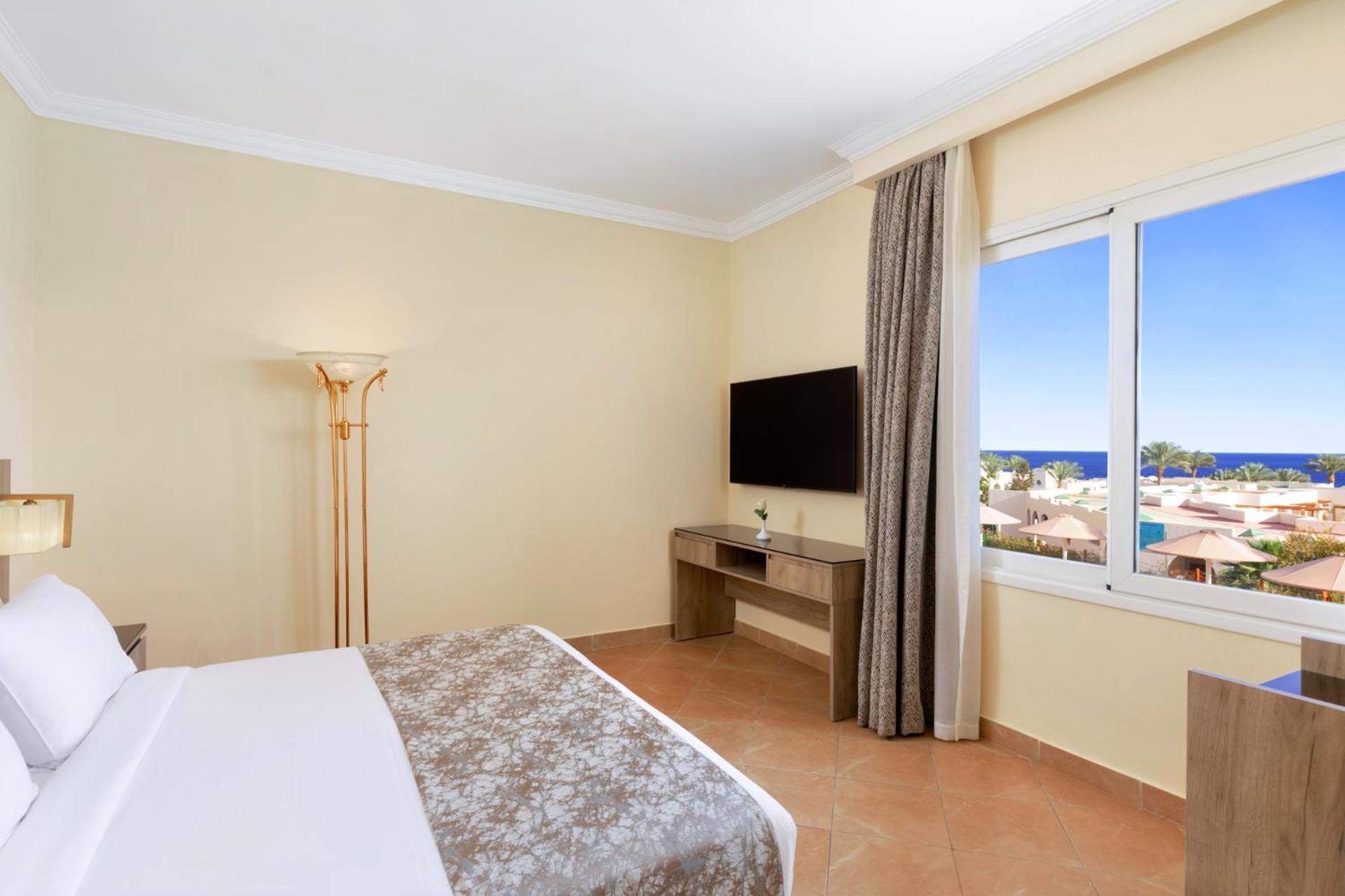 Pickalbatros Royal Grand Sharm - Adults Friendly 16 Years Plus ภายนอก รูปภาพ The photo shows a hotel room with a modern and minimalistic design. There is a comfortable bed with white linens and a decorative gray throw at the foot. A large window offers a view of the outdoors, possibly with a glimpse of the sea or beach. A fla