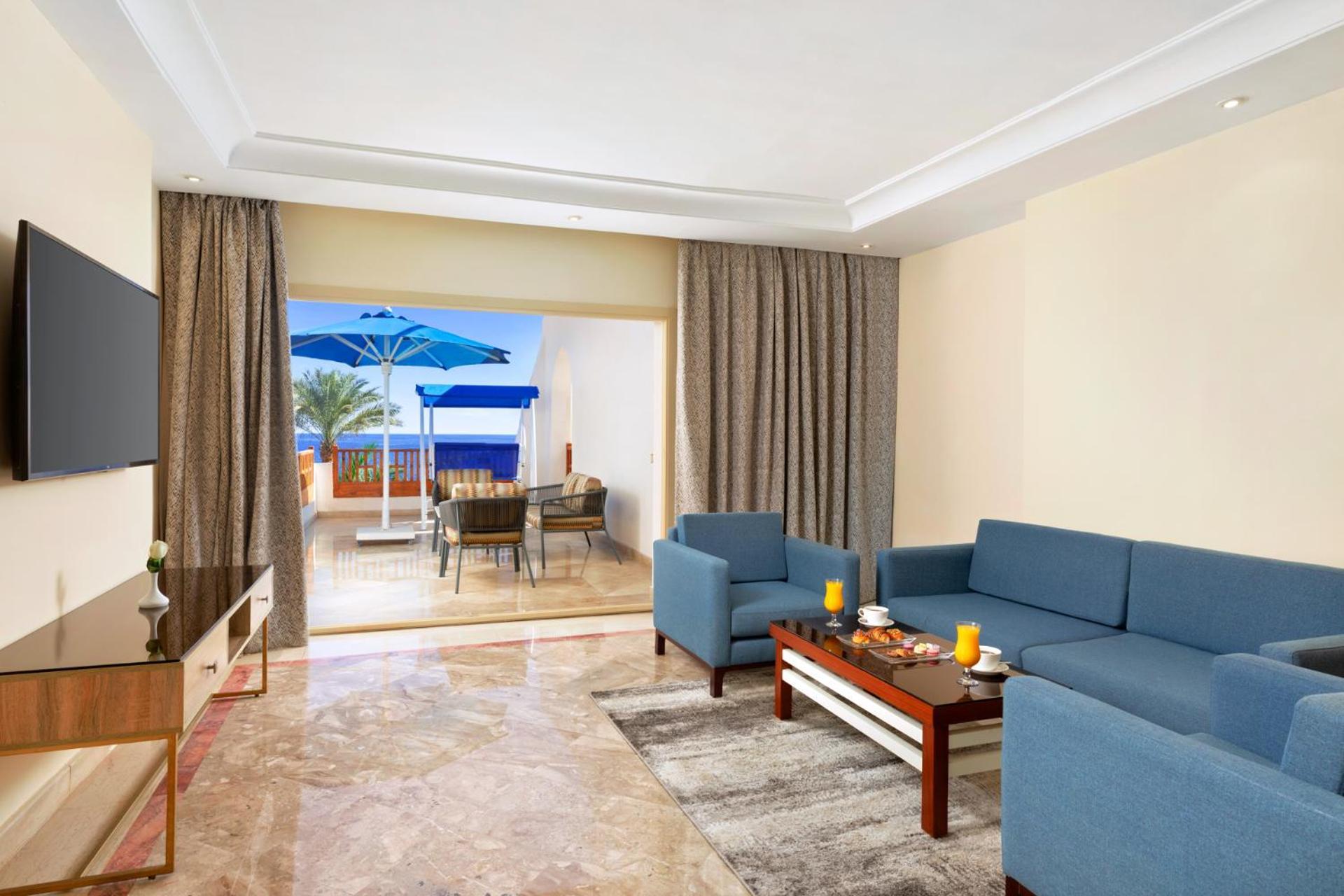 Pickalbatros Royal Grand Sharm - Adults Friendly 16 Years Plus ภายนอก รูปภาพ The photo shows a spacious living area with modern decor. There are two blue sofas arranged around a coffee table that holds some decorative items and candles. A flat-screen TV is mounted on the wall across from the seating area. To the left, there a