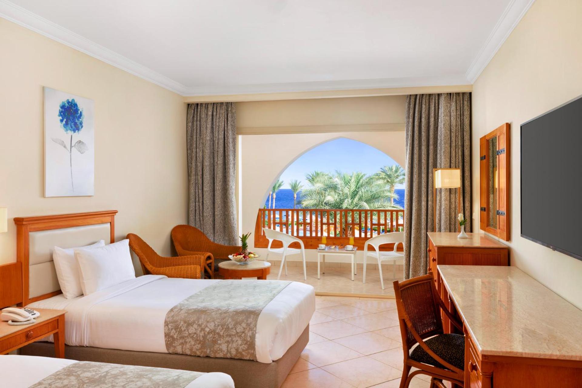Pickalbatros Royal Grand Sharm - Adults Friendly 16 Years Plus ภายนอก รูปภาพ The photo shows a hotel room featuring two beds with light bedding. There is a wooden desk and chair, and a comfortable chair is placed nearby. The room has large windows that open to a balcony with a view of palm trees and a blue sky. The decor is r