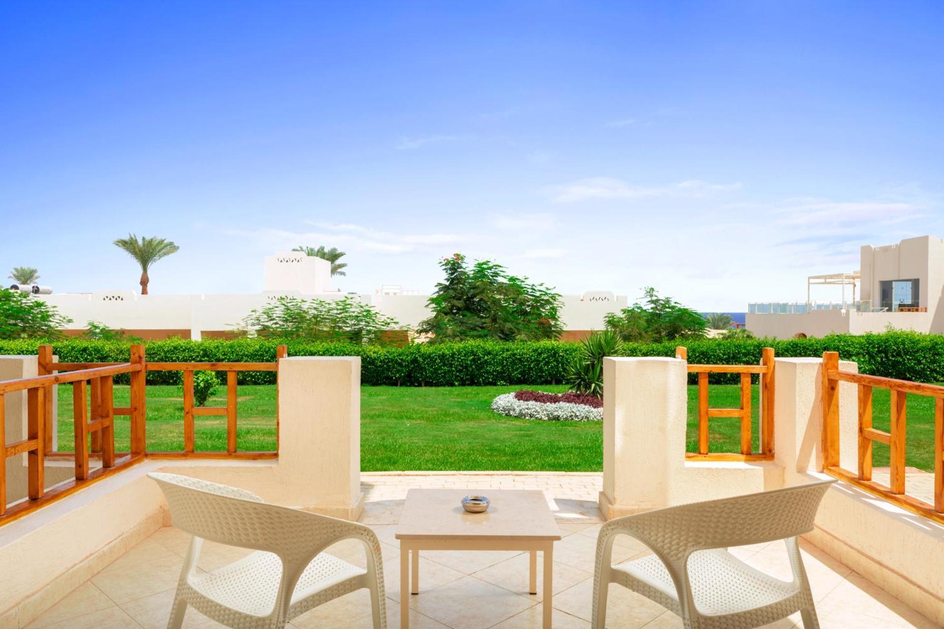 Pickalbatros Royal Grand Sharm - Adults Friendly 16 Years Plus ภายนอก รูปภาพ The photo depicts a serene outdoor balcony or patio area. It features two white chairs and a small table, set against a backdrop of lush green grass and garden plants. In the distance, there are some palm trees and clear blue skies, suggesting a warm