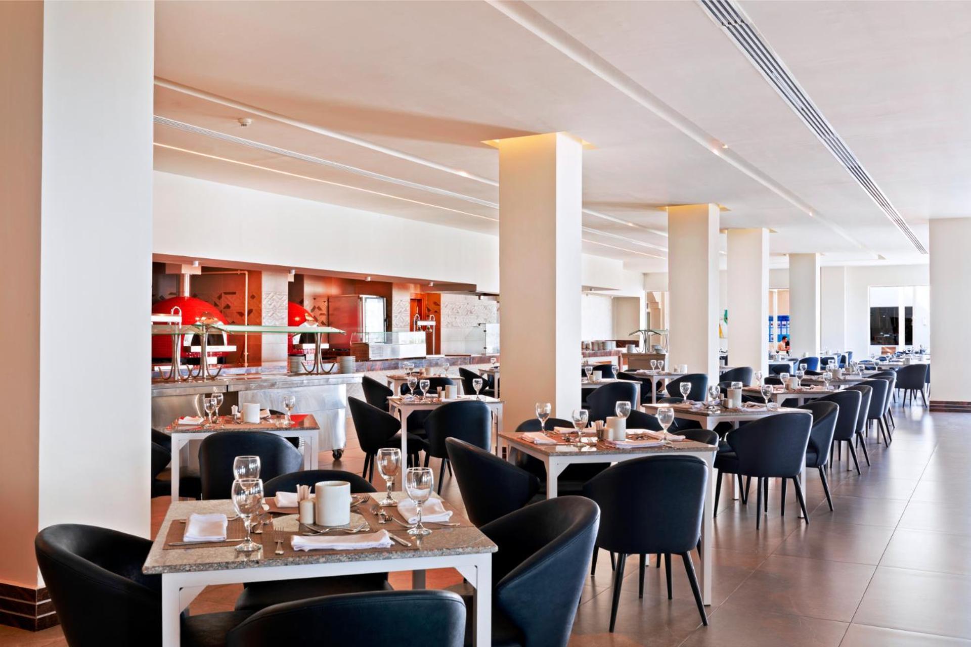 Pickalbatros Royal Grand Sharm - Adults Friendly 16 Years Plus ภายนอก รูปภาพ The photo shows a modern restaurant interior. It features a spacious layout with several dining tables arranged in a row, each set with dishes, glasses, and cutlery. The tables have light-colored surfaces and are accompanied by black chairs. In the b