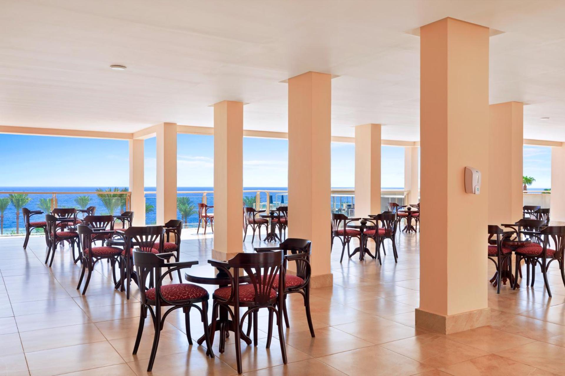 Pickalbatros Royal Grand Sharm - Adults Friendly 16 Years Plus ภายนอก รูปภาพ The image shows a dining area or café with several round wooden tables and chairs, featuring red upholstery. The space is well-lit, with large windows that allow a view of the ocean in the background, contributing to a bright and open atmosphere. The