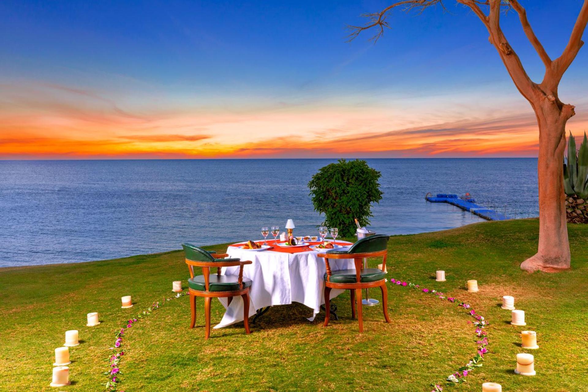 Pickalbatros Royal Grand Sharm - Adults Friendly 16 Years Plus ภายนอก รูปภาพ The photo depicts a romantic outdoor dining setup by the seaside. In the foreground, there is a round table elegantly set with a white tablecloth, plates, and glassware, creating a cozy atmosphere. Surrounding the table are green chairs. A circle of 