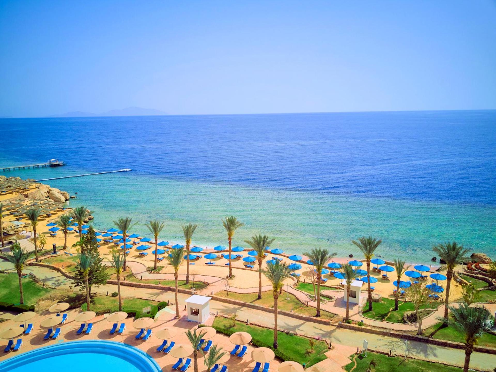 Pickalbatros Royal Grand Sharm - Adults Friendly 16 Years Plus ภายนอก รูปภาพ The photo displays a beautiful coastal scene featuring a clear blue sea that transitions into green tones near the shore. In the foreground, there are sandy beaches lined with palm trees and a variety of beach umbrellas. There is also a swimming pool