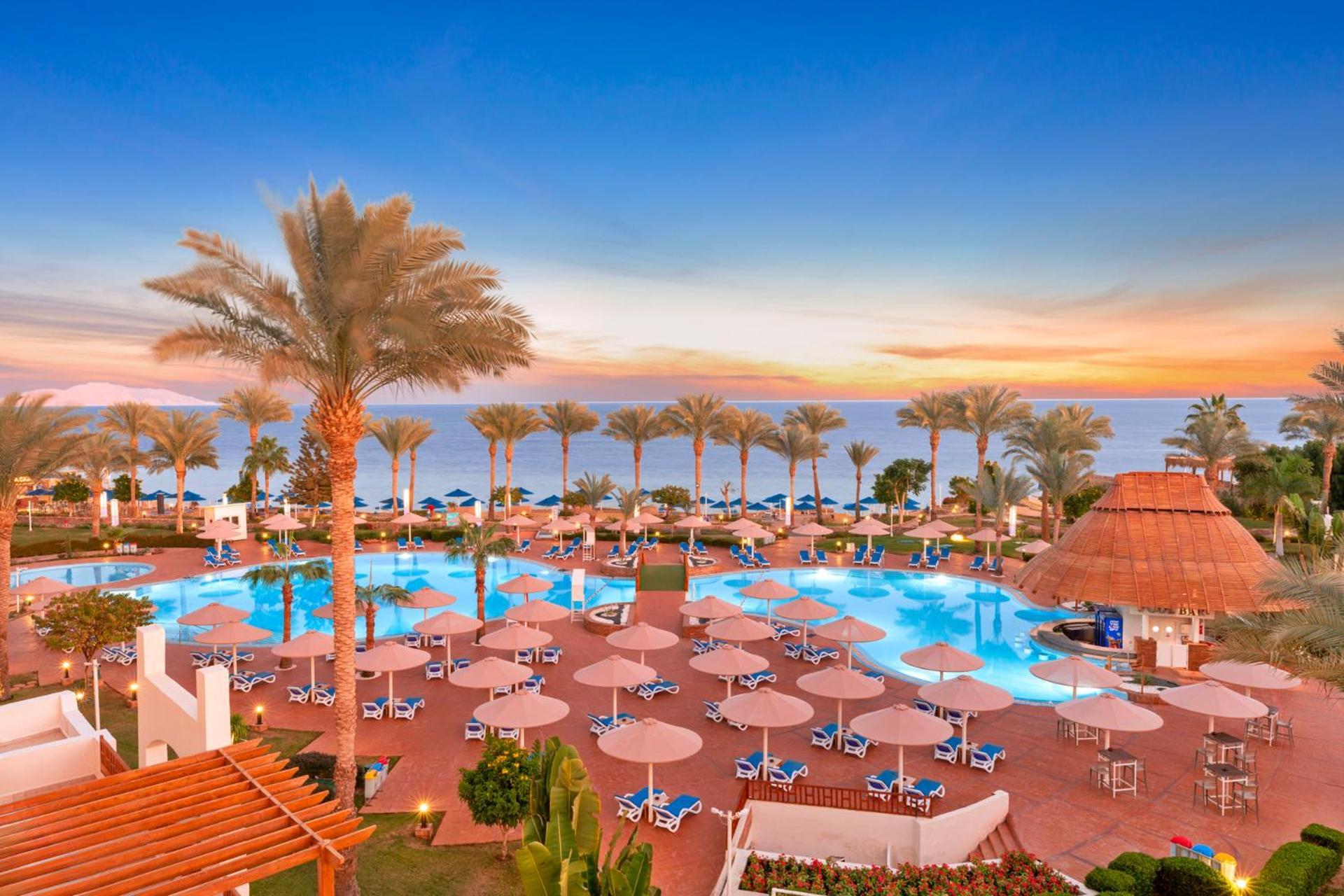 Pickalbatros Royal Grand Sharm - Adults Friendly 16 Years Plus ภายนอก รูปภาพ The photo depicts a scenic view of a resort area featuring a large swimming pool surrounded by numerous lounge chairs and umbrellas. In the background, there are tall palm trees and a beautiful sunset over the horizon, creating a vibrant sky. The atm