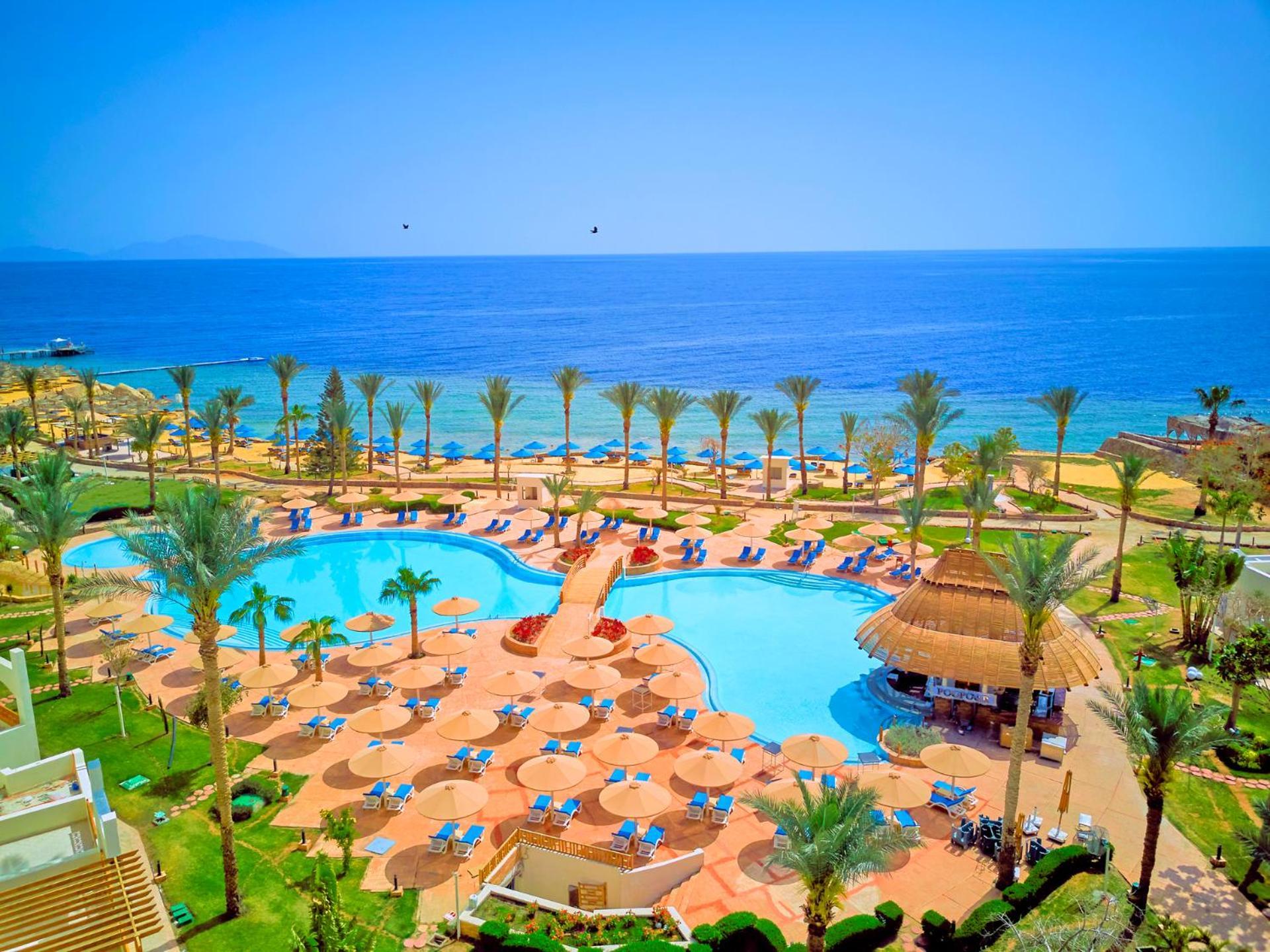 Pickalbatros Royal Grand Sharm - Adults Friendly 16 Years Plus ภายนอก รูปภาพ The photo depicts a picturesque resort scene by the sea. In the foreground, there is a large, inviting swimming pool surrounded by lounge chairs and umbrellas. Palm trees are strategically placed around the pool area, adding to the tropical atmospher