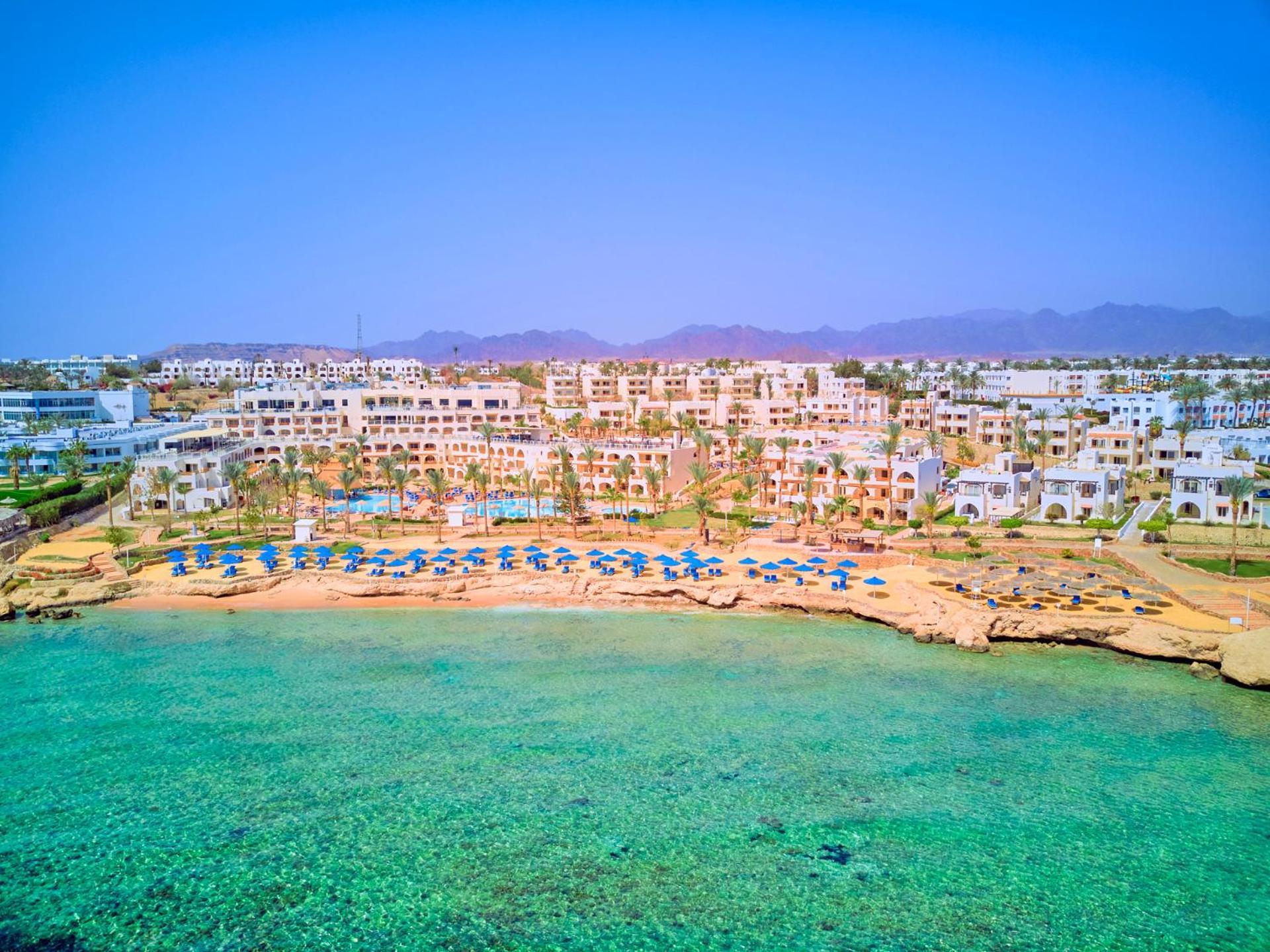 Pickalbatros Royal Grand Sharm - Adults Friendly 16 Years Plus ภายนอก รูปภาพ The photo depicts a picturesque beachfront resort. It shows a clear turquoise sea with a sandy shore lined with beach umbrellas and sun loungers. Behind the beach, there are several white buildings of the resort, surrounded by lush greenery. In the b