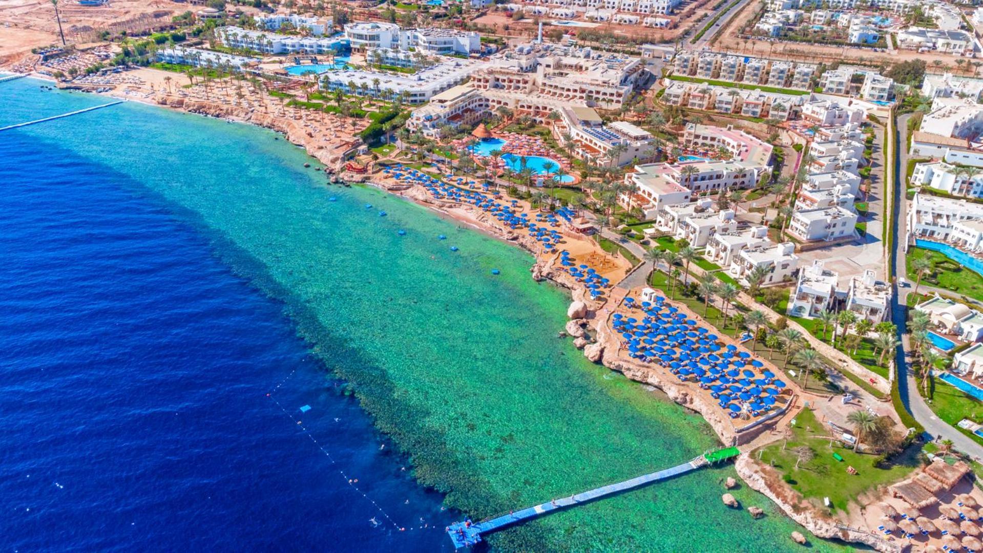Pickalbatros Royal Grand Sharm - Adults Friendly 16 Years Plus ภายนอก รูปภาพ The photo showcases a beautiful aerial view of a coastal resort. In the foreground, there's a sandy beach with beach umbrellas and lounge chairs arranged neatly, indicating a tourist area. The clear, turquoise water transitions from the sandy shore t
