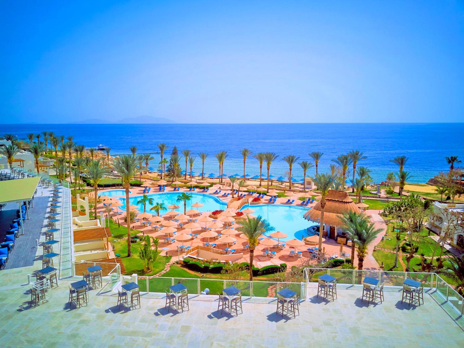 Pickalbatros Royal Grand Sharm - Adults Friendly 16 Years Plus ภายนอก รูปภาพ The photo shows a beautiful resort scene with a spacious swimming pool at the center, surrounded by sun loungers and palm trees. There are areas for relaxation with tables and chairs positioned around the pool. In the background, the vibrant blue oce