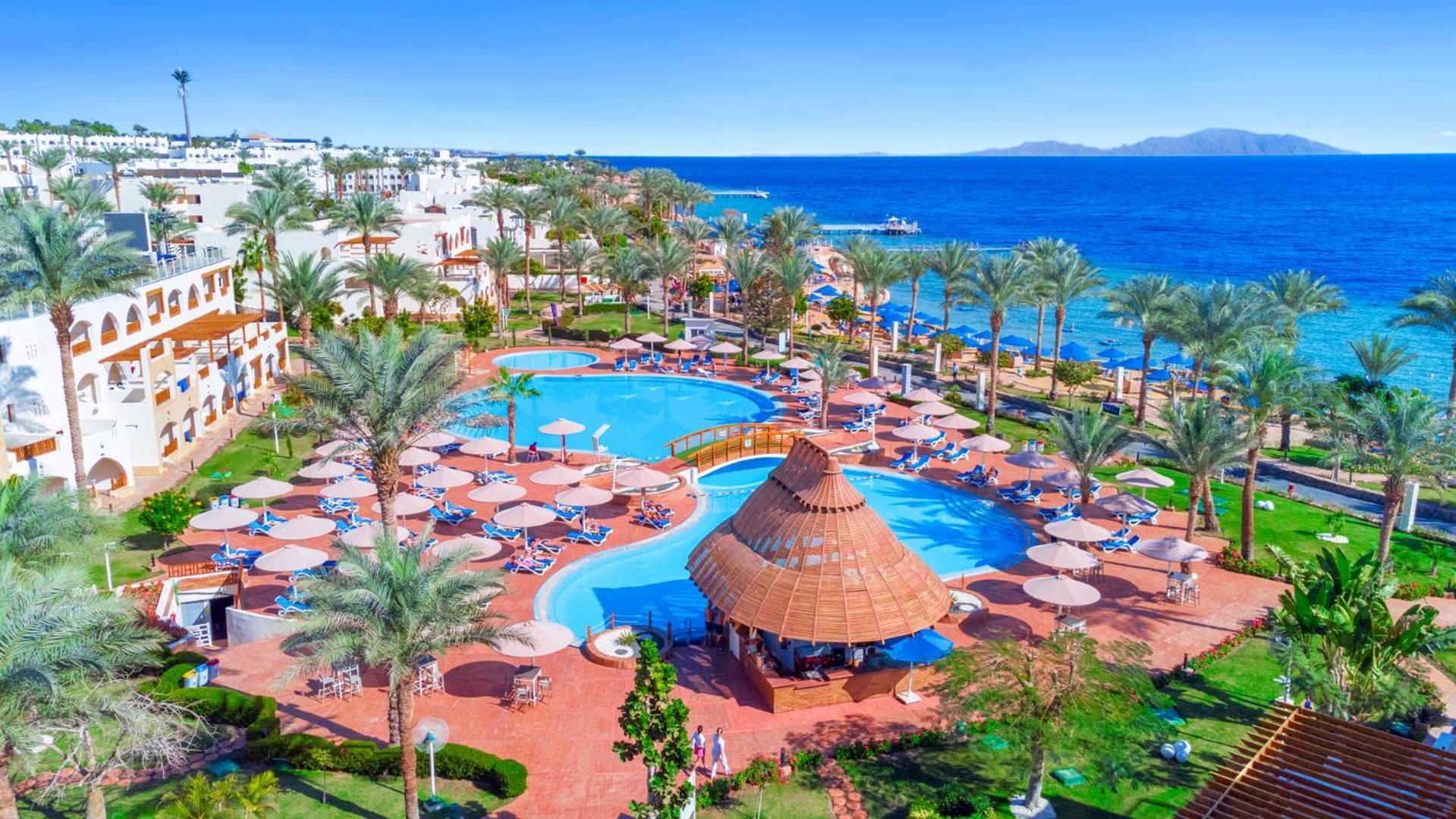 Pickalbatros Royal Grand Sharm - Adults Friendly 16 Years Plus ภายนอก รูปภาพ The image depicts a vibrant resort scene overlooking a clear blue sea. In the foreground, there are multiple swimming pools surrounded by sun loungers and umbrellas. A thatched-roof gazebo is situated near the center of the pool area. Palms and lush 