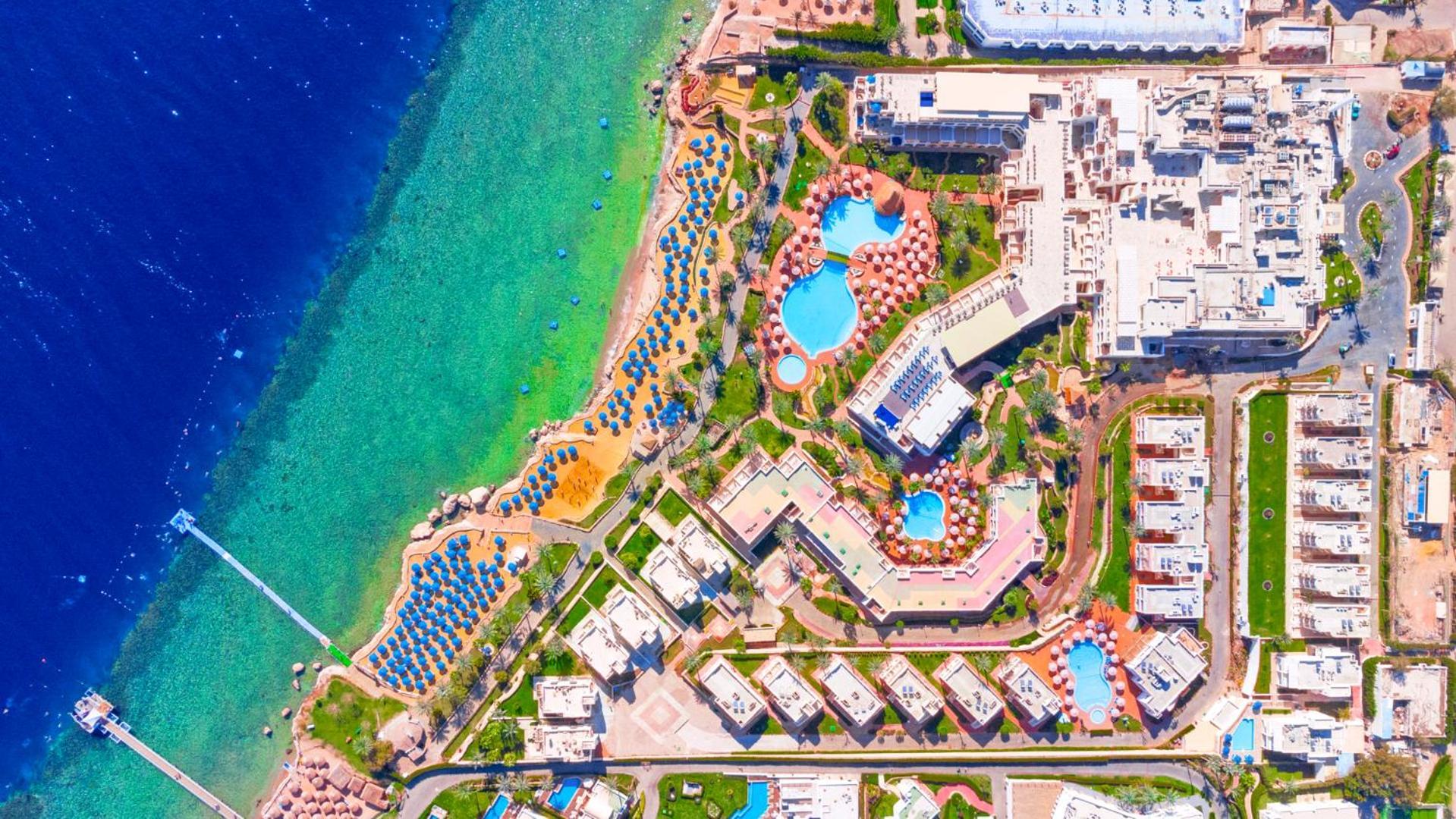 Pickalbatros Royal Grand Sharm - Adults Friendly 16 Years Plus ภายนอก รูปภาพ The image shows an aerial view of a coastal resort area. It features various swimming pools outlined by landscaped gardens and pathways. There are also rows of sun loungers and umbrellas near the beach. The water transitions from a deep blue to a vib