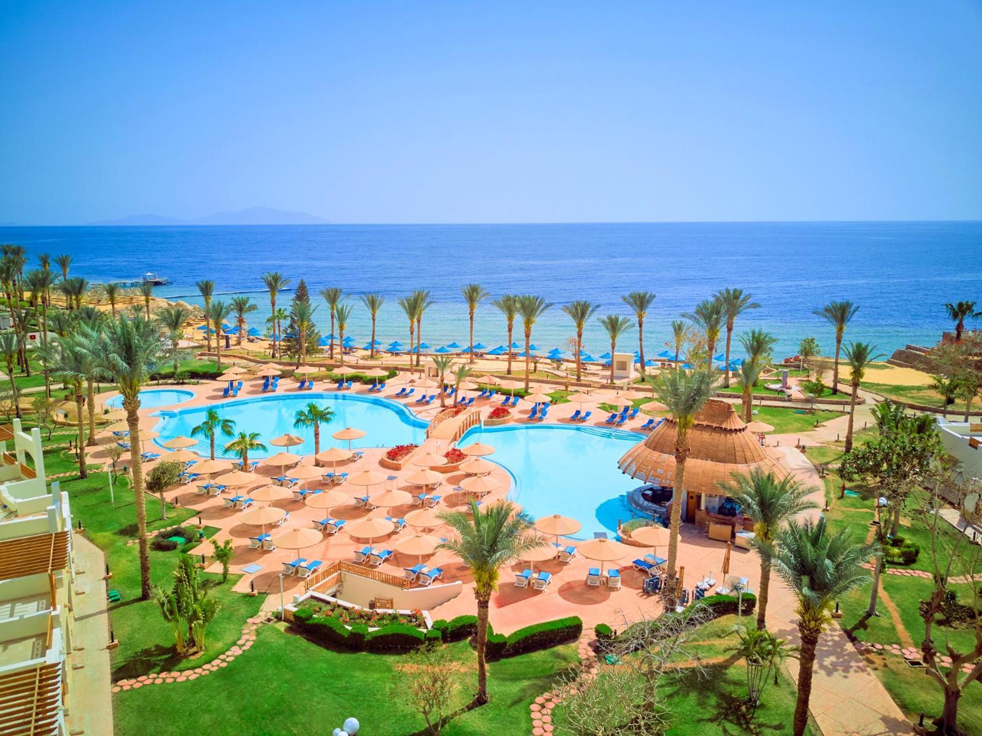 Pickalbatros Royal Grand Sharm - Adults Friendly 16 Years Plus ภายนอก รูปภาพ The photo depicts a beautiful resort scene by the ocean. In the foreground, there is a large swimming pool surrounded by palm trees and lounge chairs. The pool area features sunbeds and umbrellas, suggesting a relaxing atmosphere for guests. In the b