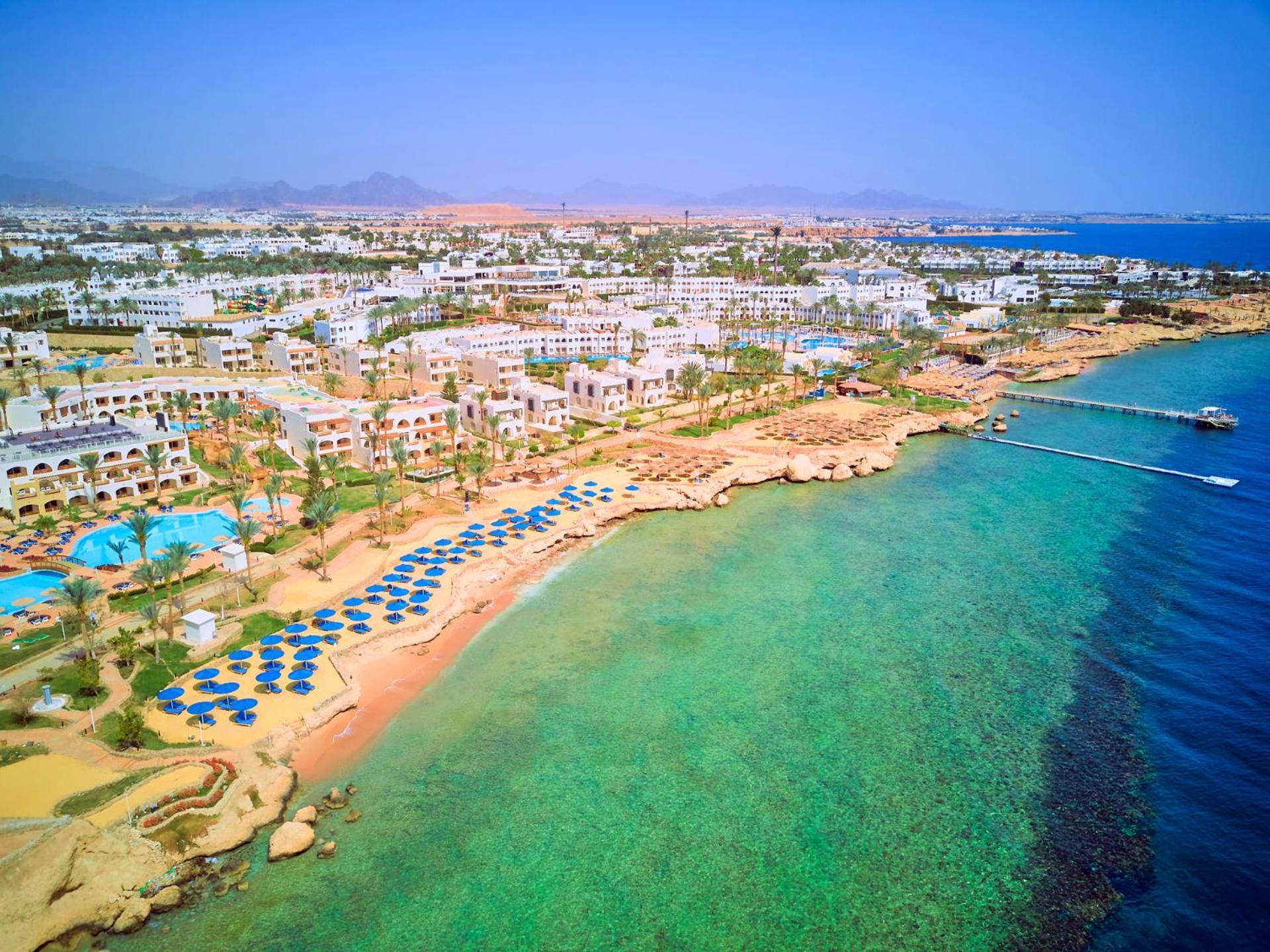 Pickalbatros Royal Grand Sharm - Adults Friendly 16 Years Plus ภายนอก รูปภาพ The photo showcases a picturesque coastal scene, likely from a resort area. It features a sandy beach lined with blue umbrellas and lounge chairs, indicating a recreational space for relaxation. The water is clear and exhibits shades of green and blu