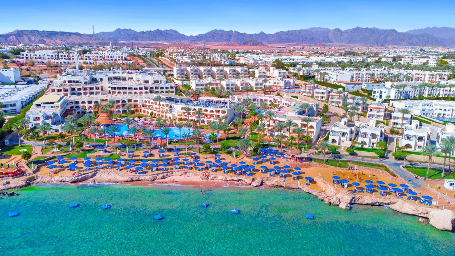Pickalbatros Royal Grand Sharm - Adults Friendly 16 Years Plus ภายนอก รูปภาพ The photo showcases a beautiful coastal resort area. In the foreground, there is a sandy beach lined with blue umbrellas and sun loungers. The vibrant turquoise water gently laps at the shore. Behind the beach, you can see a well-maintained resort wi
