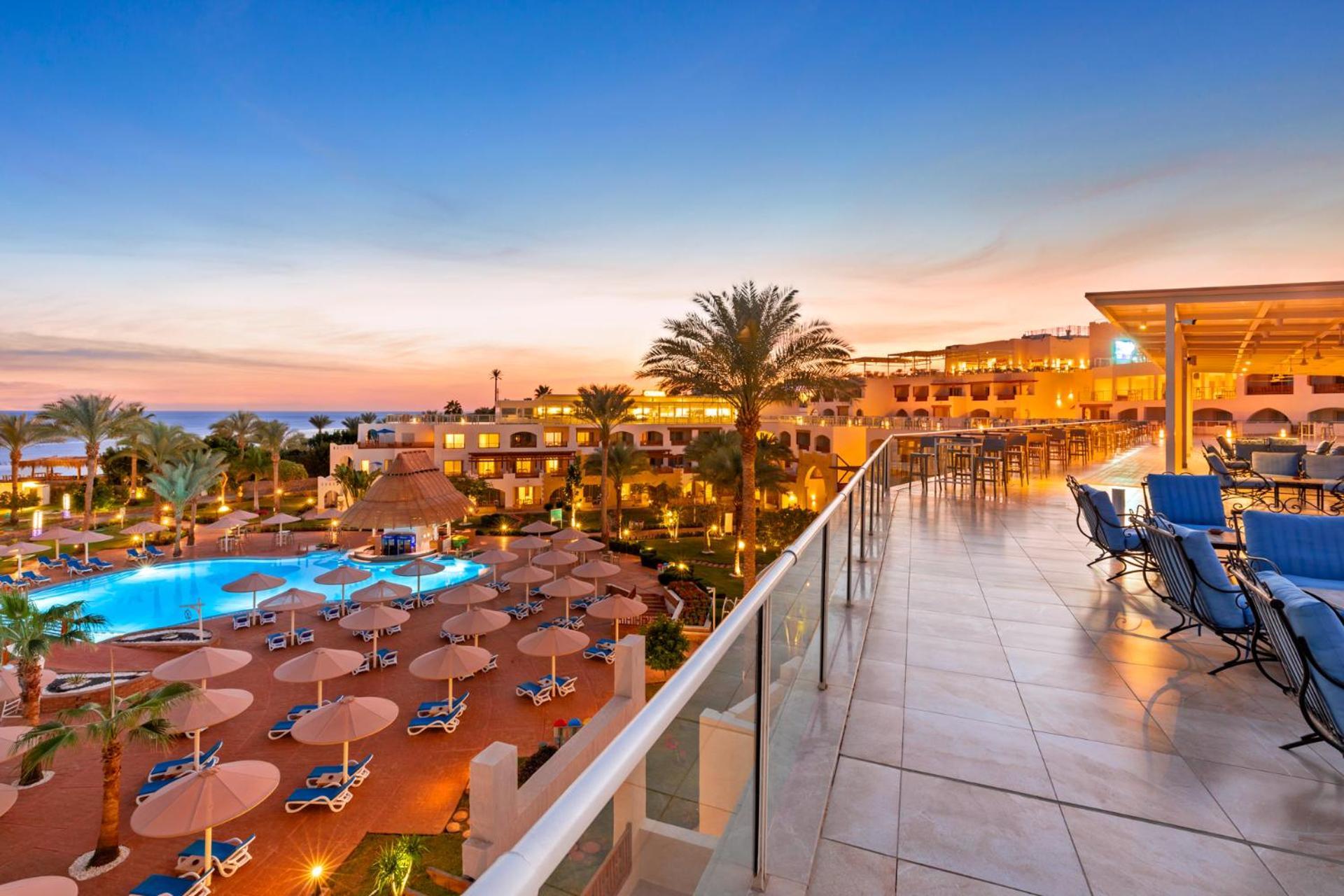 Pickalbatros Royal Grand Sharm - Adults Friendly 16 Years Plus ภายนอก รูปภาพ The photo depicts a scenic view of a resort during sunset. In the foreground, there is a balcony with tiled flooring, overlooking the resort. Below, a swimming pool is visible, surrounded by lounge chairs and large umbrellas, indicating a relaxing at