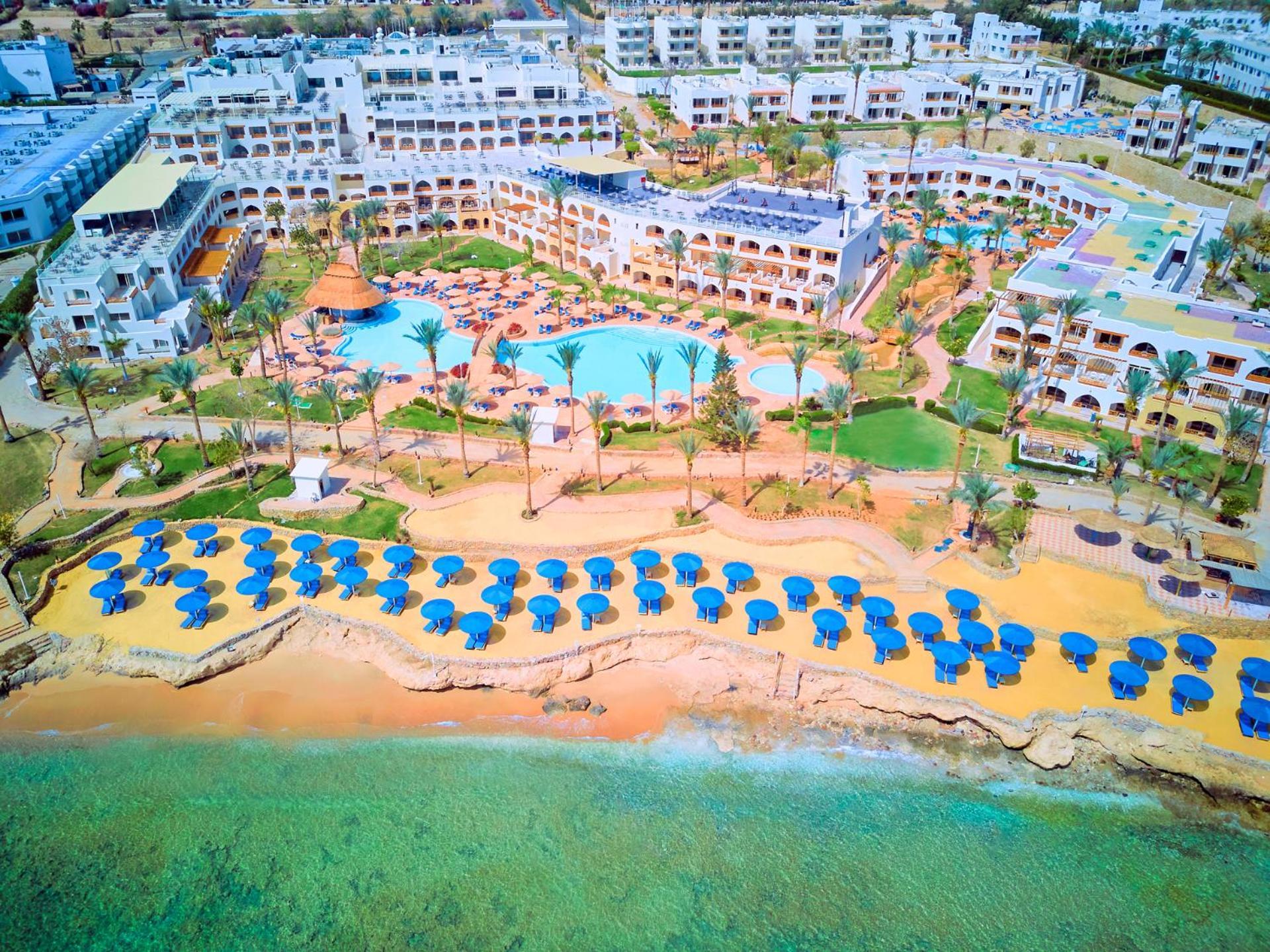 Pickalbatros Royal Grand Sharm - Adults Friendly 16 Years Plus ภายนอก รูปภาพ The photo shows a large resort complex situated near a beach. There is a turquoise swimming pool at the center, surrounded by green lawns and palm trees. Sun loungers with blue umbrellas are lined up on the sandy beach, and the beach area is adjacent