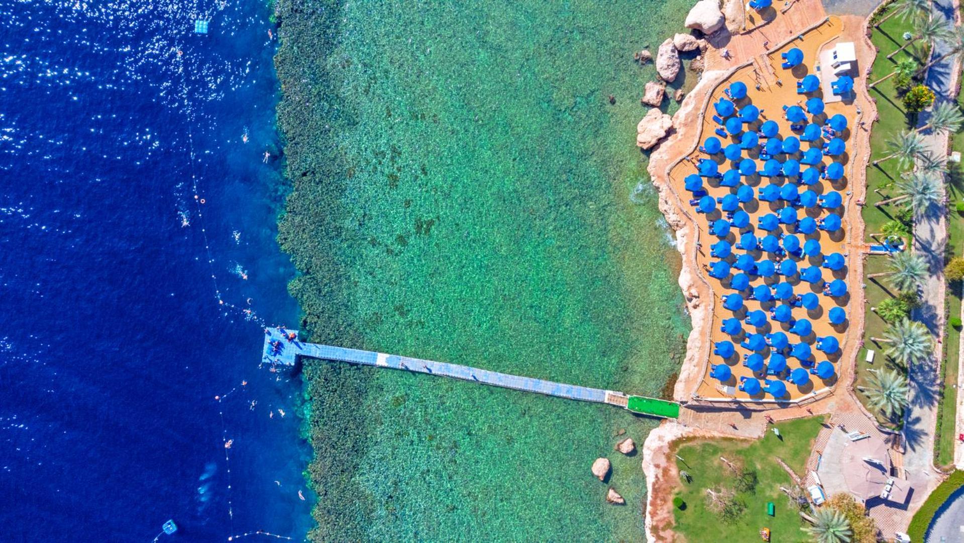 Pickalbatros Royal Grand Sharm - Adults Friendly 16 Years Plus ภายนอก รูปภาพ The photo shows an aerial view of a beach area. In the foreground, there is a wooden pier extending into the clear blue water. To the right, there are numerous beach umbrellas with blue canopies arranged neatly, likely indicating a resort or beach cl