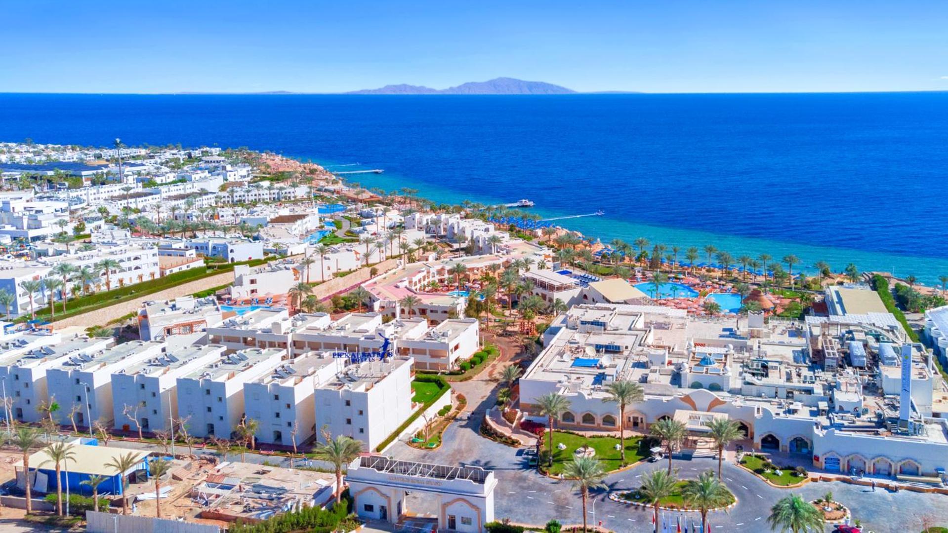 Pickalbatros Royal Grand Sharm - Adults Friendly 16 Years Plus ภายนอก รูปภาพ The photo shows a beautiful coastal landscape, featuring a resort area by the sea. There are white buildings, likely hotels or villas, arranged in a sprawling layout near the beach. The water is a vibrant blue, contrasting with the sandy shore and gr