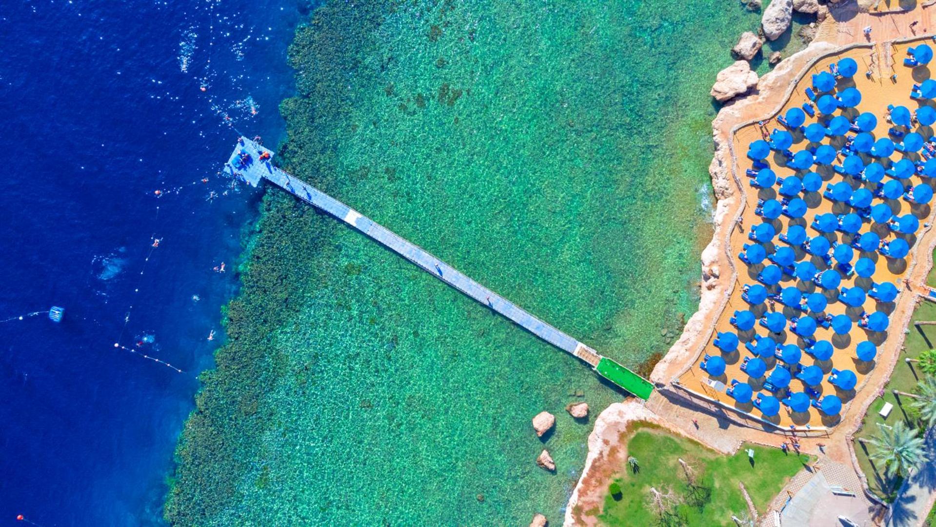 Pickalbatros Royal Grand Sharm - Adults Friendly 16 Years Plus ภายนอก รูปภาพ The photo shows an aerial view of a coastal area featuring a long wooden pier extending into clear turquoise water. On one side of the pier, there appears to be a beach with umbrellas or sunbeds arranged neatly. The water is vibrant and visually invi