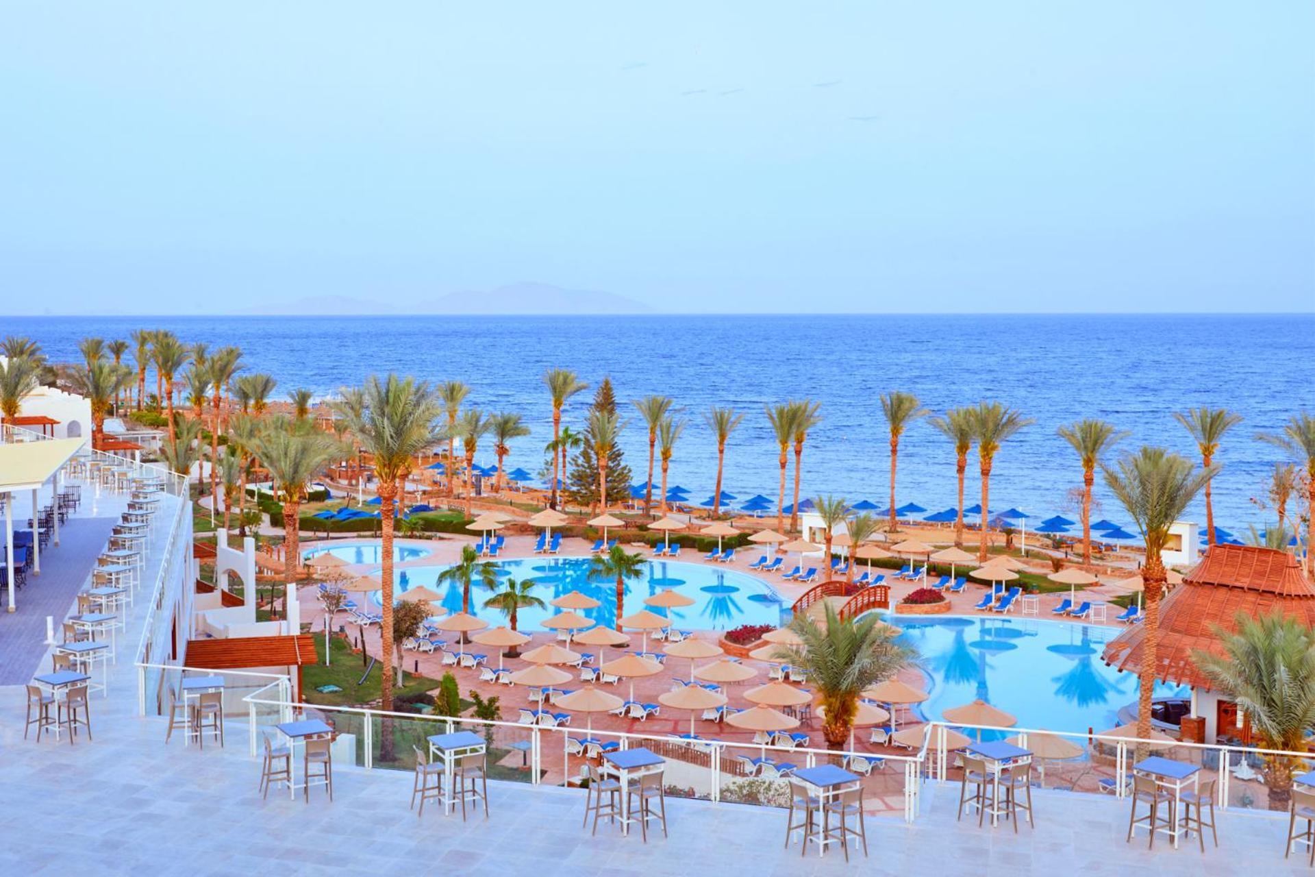 Pickalbatros Royal Grand Sharm - Adults Friendly 16 Years Plus ภายนอก รูปภาพ The photo depicts a scenic resort area by the sea. In the foreground, there is a large swimming pool surrounded by lounge chairs and umbrellas. Palm trees line the sides of the pool, adding to the tropical atmosphere. In the background, a beautiful b