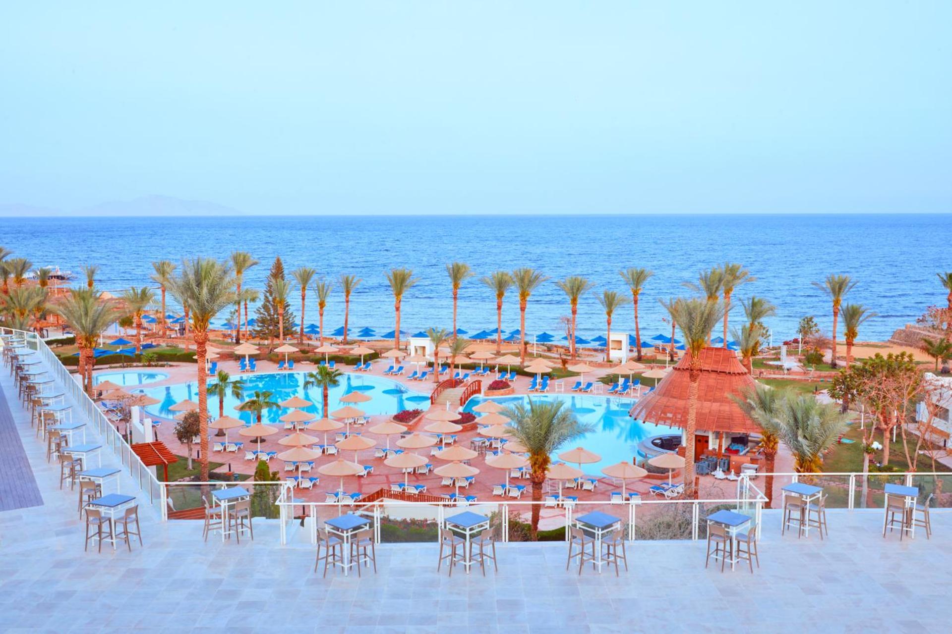 Pickalbatros Royal Grand Sharm - Adults Friendly 16 Years Plus ภายนอก รูปภาพ The photo shows a scenic view of a resort or beach area. In the foreground, there are several seating areas with bar stools on a terrace. Below, a large swimming pool is visible, surrounded by lounging chairs and umbrellas. Palm trees line the area, 