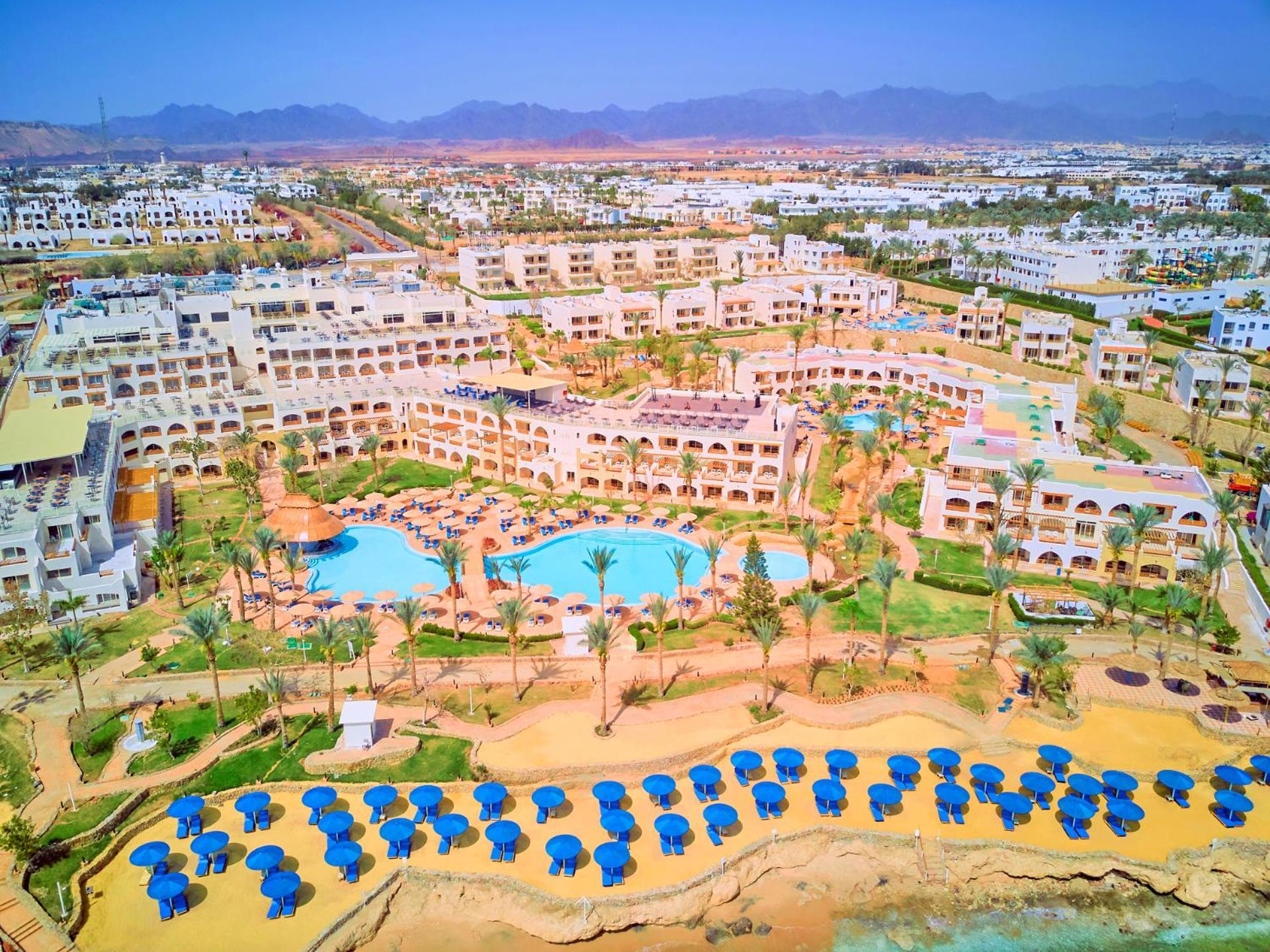 Pickalbatros Royal Grand Sharm - Adults Friendly 16 Years Plus ภายนอก รูปภาพ The photo shows a large resort complex set in a scenic location, likely near a beach. The layout features multiple buildings with a Mediterranean style of architecture, surrounded by lush greenery and palm trees. Central to the resort is a swimming p