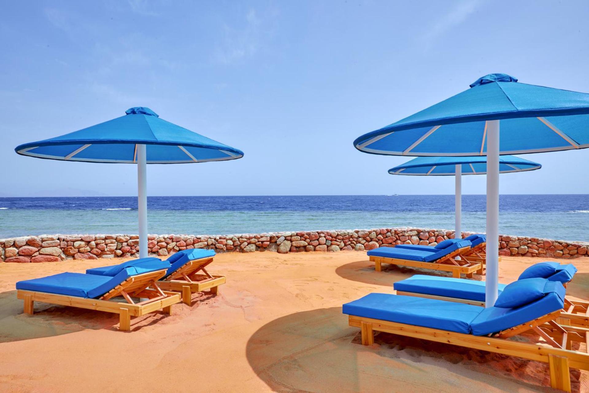 Pickalbatros Royal Grand Sharm - Adults Friendly 16 Years Plus ภายนอก รูปภาพ The photo shows a beach scene featuring several blue umbrellas and lounge chairs on a sandy area. The umbrellas are large and have a bright blue color, providing shade for the lounge chairs underneath. In the background, there's a view of the ocean w