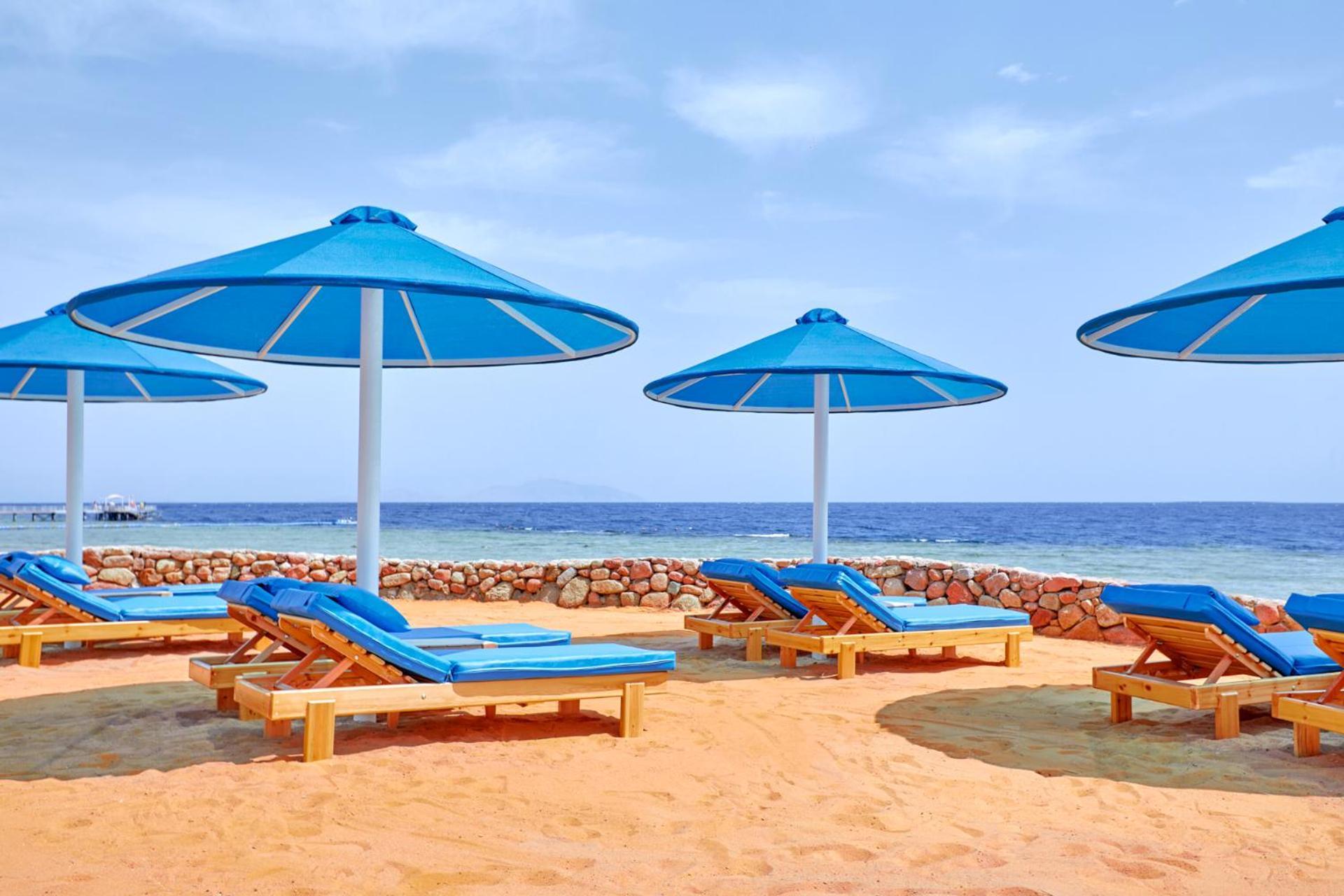 Pickalbatros Royal Grand Sharm - Adults Friendly 16 Years Plus ภายนอก รูปภาพ The photo shows a beach scene featuring several blue umbrellas providing shade over wooden lounge chairs. The sandy beach extends toward a calm sea, creating a relaxing atmosphere. The sky is mostly clear, with a few clouds, suggesting pleasant weath