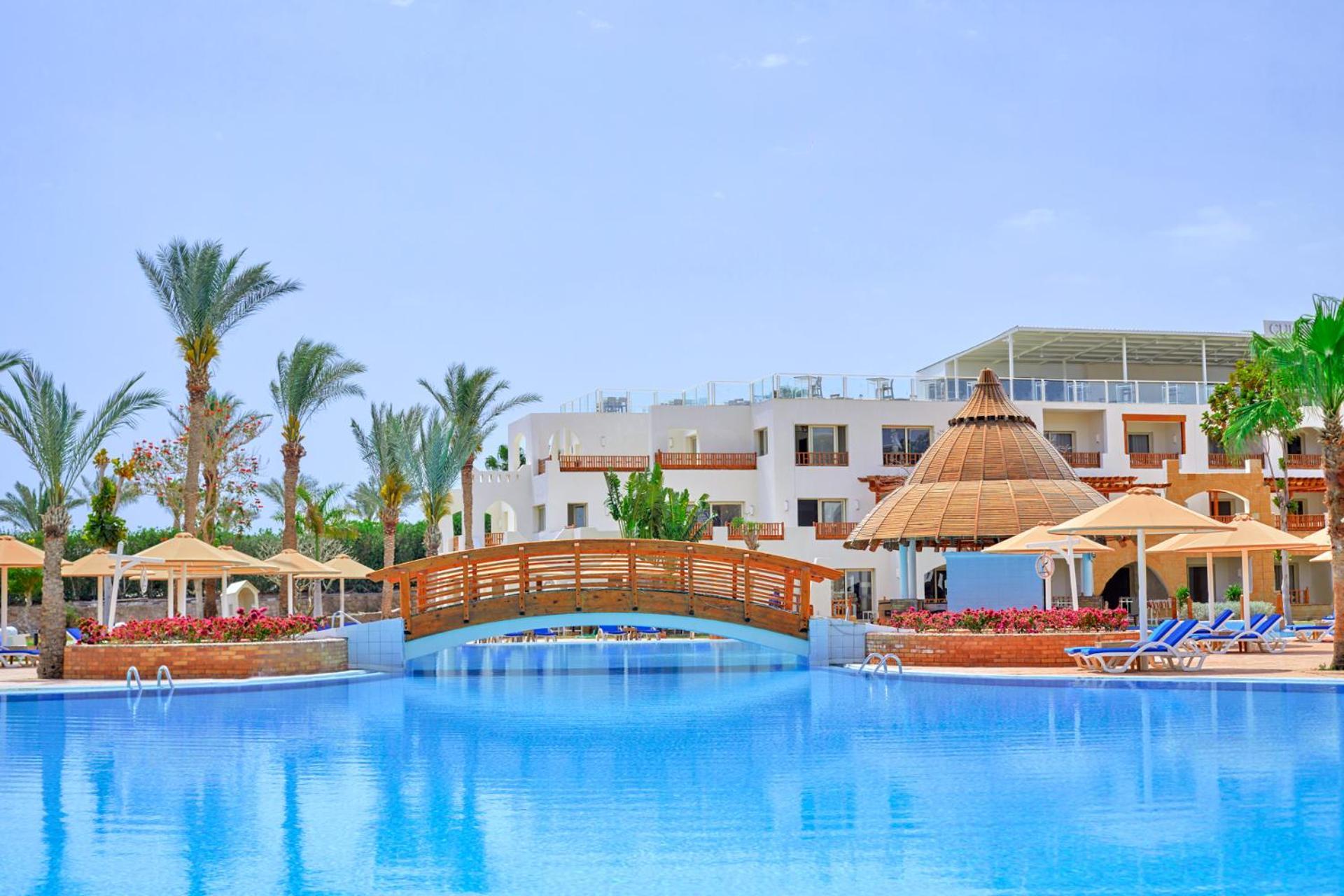 Pickalbatros Royal Grand Sharm - Adults Friendly 16 Years Plus ภายนอก รูปภาพ The photo depicts a serene resort setting featuring a large, calm swimming pool, which reflects the clear blue sky. In the foreground, a wooden bridge arches over the pool. Surrounding the pool are several palm trees and tropical foliage, contributin