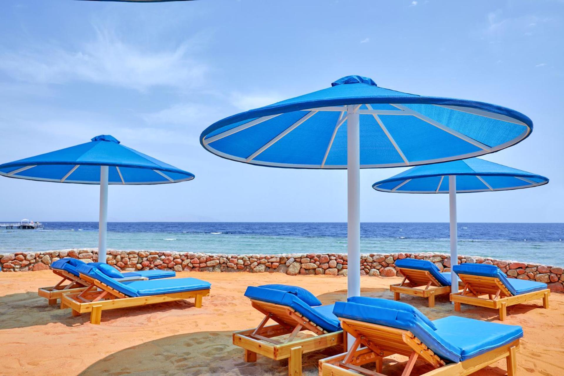 Pickalbatros Royal Grand Sharm - Adults Friendly 16 Years Plus ภายนอก รูปภาพ The photo shows a serene beach scene with several blue sun loungers arranged beneath large blue umbrellas. The loungers are positioned on sandy ground, with the ocean visible in the background. The sky is clear, indicating a bright and sunny day, and