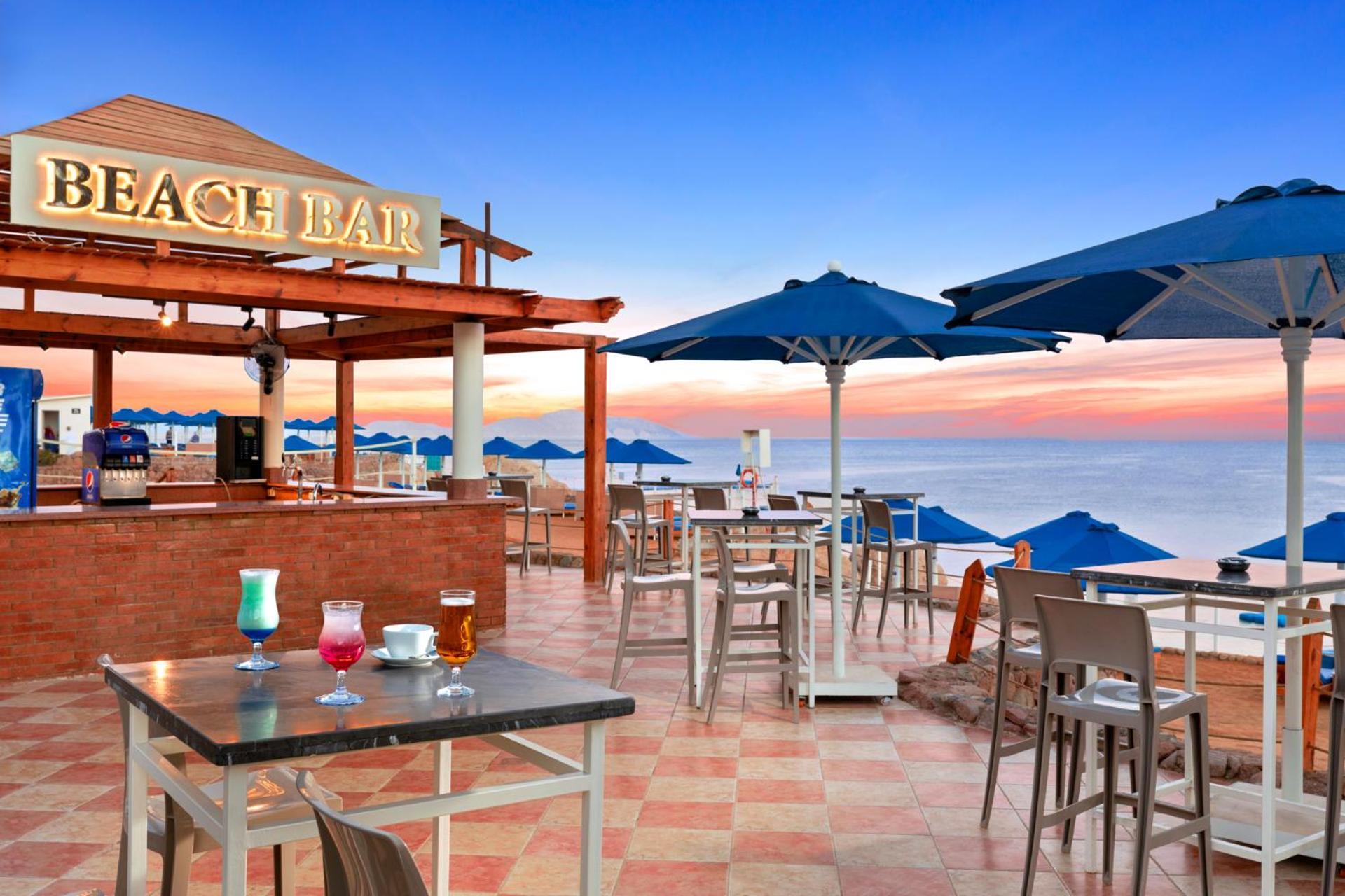 Pickalbatros Royal Grand Sharm - Adults Friendly 16 Years Plus ภายนอก รูปภาพ The photo shows a beach bar setting, featuring a wooden structure with the words "BEACH BAR" prominently displayed. There are several tables and chairs arranged on a tiled floor, with some drinks placed on the tables. Blue umbrellas provide shade for