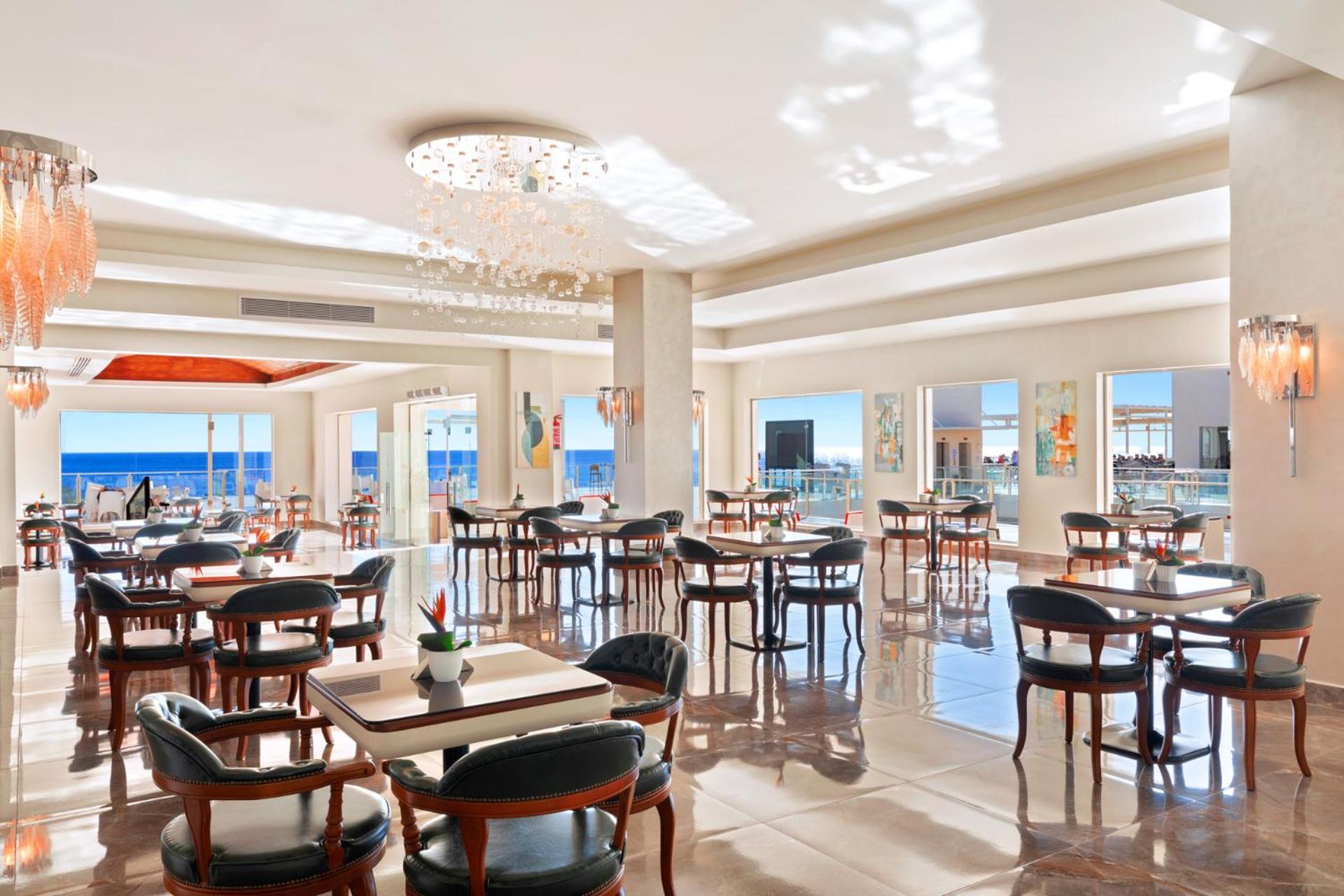Pickalbatros Royal Grand Sharm - Adults Friendly 16 Years Plus ภายนอก รูปภาพ The photo shows a bright and elegant restaurant interior. There are several tables arranged throughout the space, each with chairs around them. The decor features modern lighting, including a chandelier, and large windows that allow natural light to 