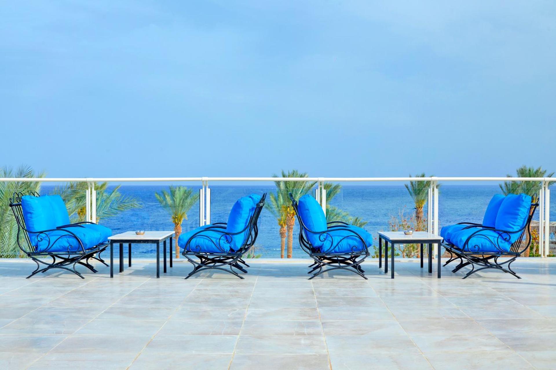 Pickalbatros Royal Grand Sharm - Adults Friendly 16 Years Plus ภายนอก รูปภาพ The photo depicts a serene outdoor patio area with four blue lounge chairs arranged around small tables. The setting features a clear view of the ocean with palm trees visible in the background, complementing the tranquil atmosphere. The overall colo